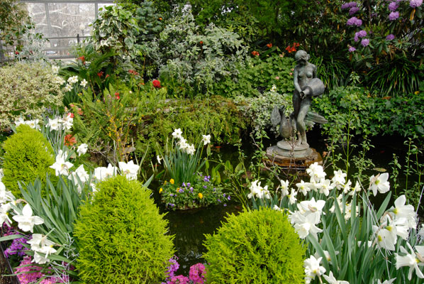 a sculpture of a woman carrying an urn at the edge of a pond inside the conservatory